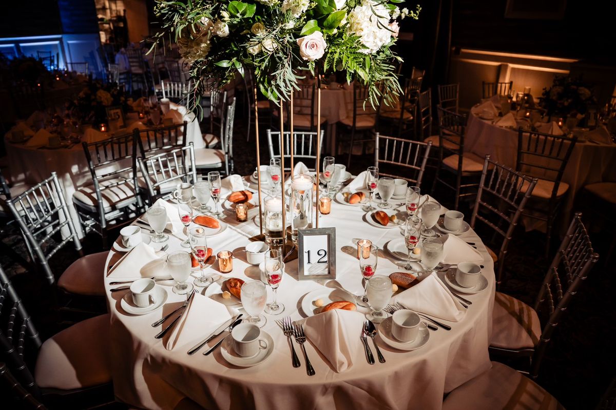 A reception table set for dinner beneath a tall floral centerpiece, with candles and champagne flutes